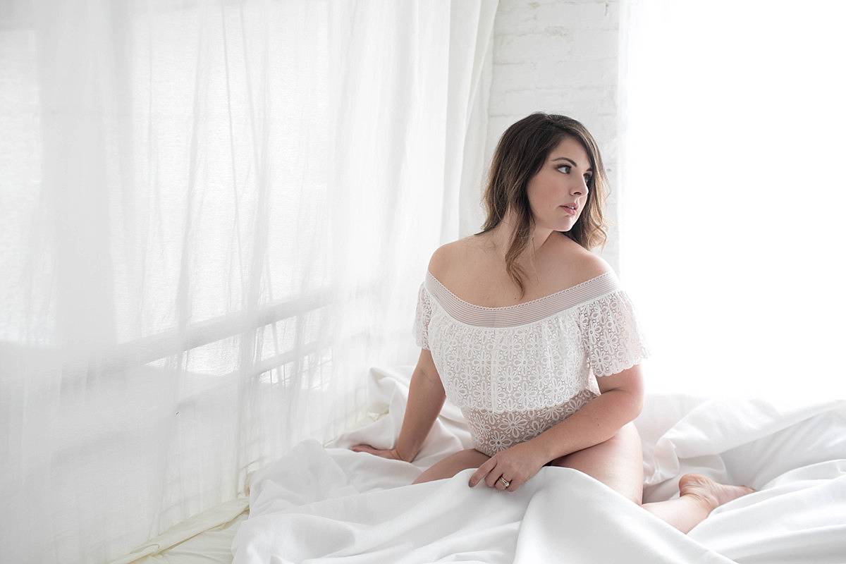 a woman on a bed in a white body suite for her boudoir photoshoot